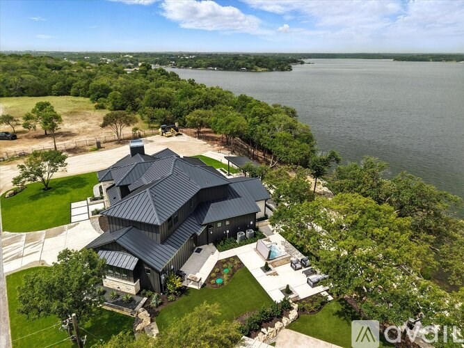 A modern house with a black roof is surrounded by greenery and overlooks a large body of water.