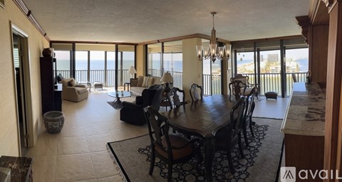 A living room with a dining table and chairs, a sofa, a chandelier, and a view of the ocean through the windows.