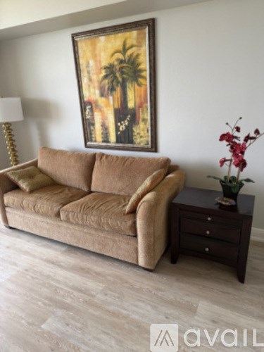 A living room with a brown couch, a lamp, a painting, and a small table with a vase of flowers.
