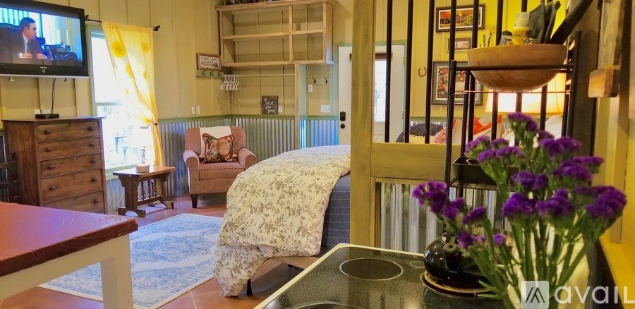 A living room with a TV, a couch, a chair, a table, a vase with purple flowers, and a shelf with a bowl on it.