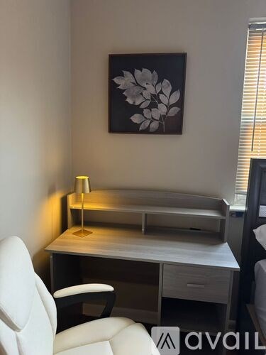 A white chair is in front of a desk with a lamp and a picture on the wall.