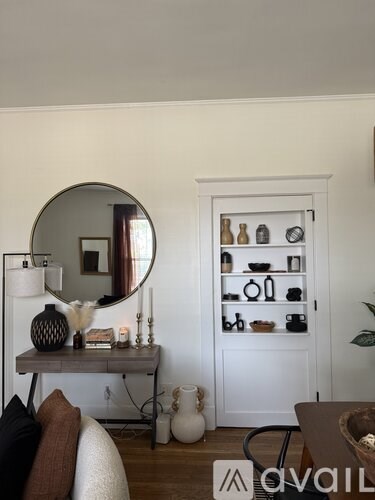 A room with a round mirror, a table with a vase and candles, and a shelf with various items.