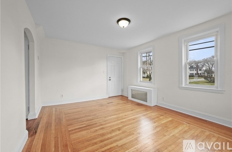 A room with wooden flooring and white walls, with a window showing a view of trees and houses outside.