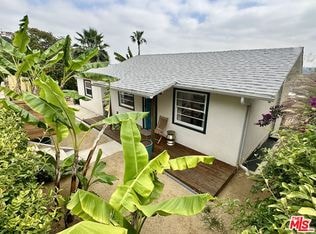 A small white house with a grey roof surrounded by green plants.