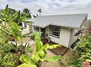 A small white house with a grey roof surrounded by green plants.
