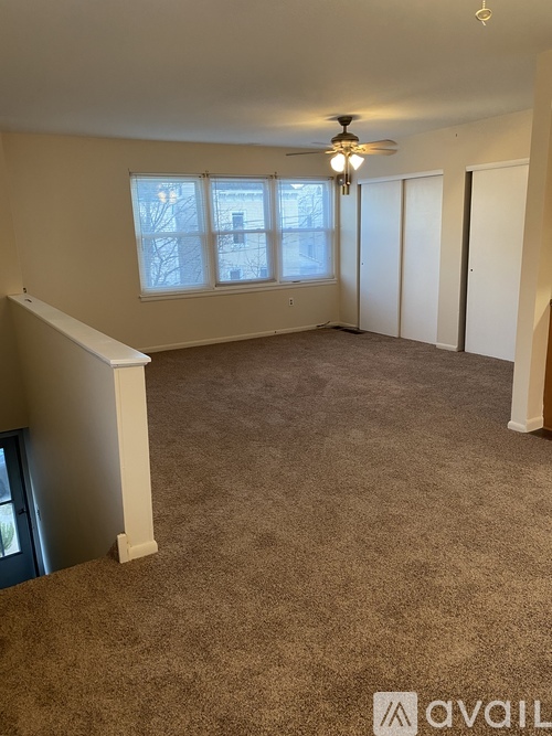 A room with a carpeted floor, a ceiling fan, and a window with a view of a building outside.