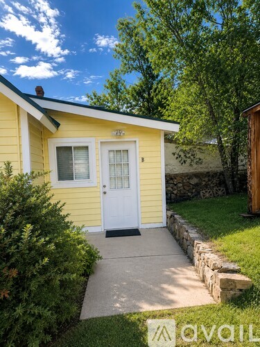 A small yellow house with a white door and windows is surrounded by greenery and a stone wall.