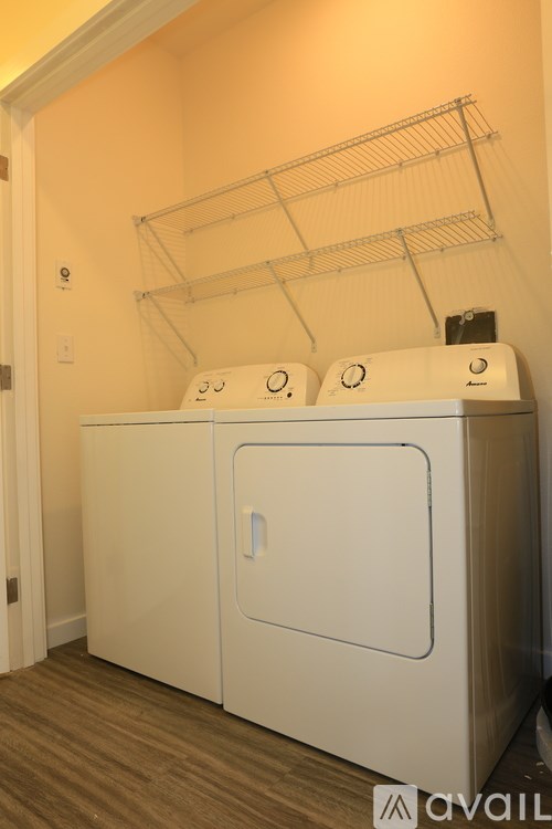 A white washing machine with a dryer on top is in a room with a white wall and wooden floor.