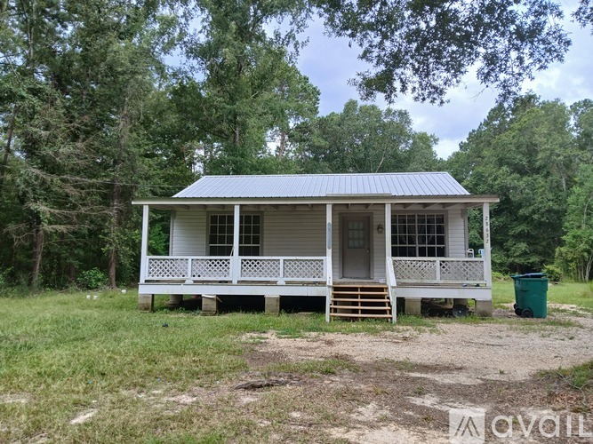 A small white house with a porch and a blue roof is surrounded by trees.