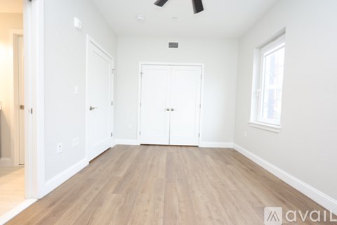 A room with white walls and wooden flooring, featuring a ceiling fan and a window.