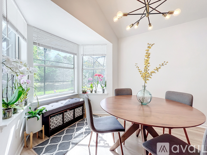 A dining room with a round table and chairs, a vase with flowers, and a window with blinds.
