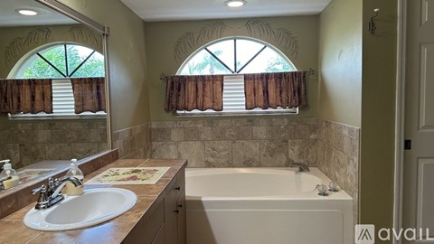 A bathroom with a sink, tub, and two windows with curtains.