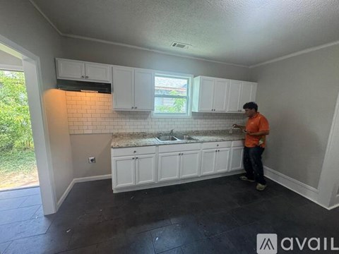 A man in an orange shirt is standing in a kitchen with white cabinets and a tiled backsplash.