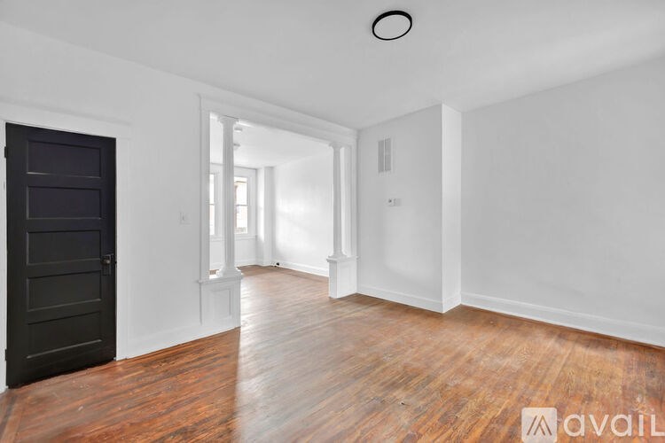 A room with wooden floors and white walls, with a black door on the left and a window with white curtains on the right.