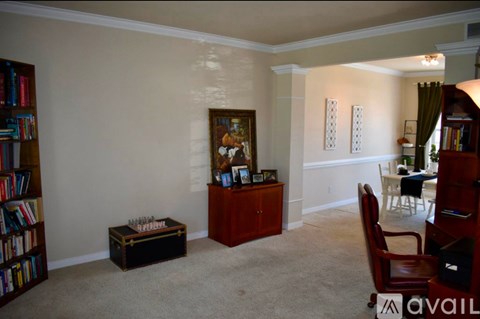 A living room with a brown chair, a brown cabinet, a brown bookcase, a painting, a small table with a vase and a candle on it, and a window with curtains.