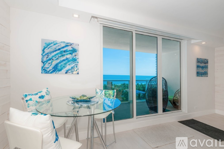 A dining room with a glass table and chairs, a painting on the wall, and a view of the outdoors through the sliding glass doors.