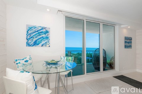 A dining room with a glass table and chairs, a painting on the wall, and a view of the outdoors through the sliding glass doors.