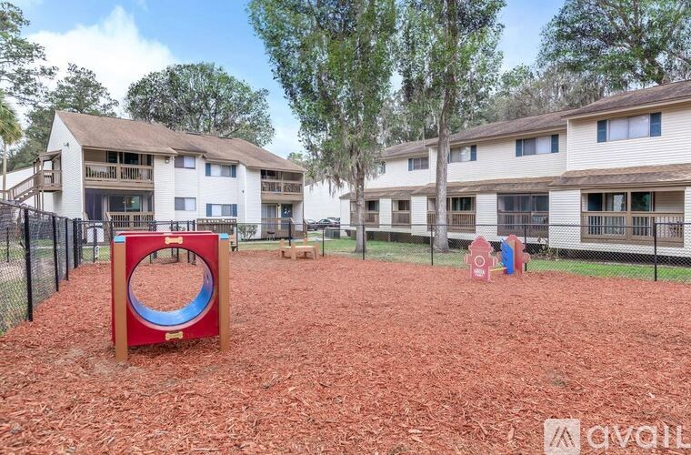 A playground area with a red and blue play structure in the middle of a dirt area with apartment buildings in the background.