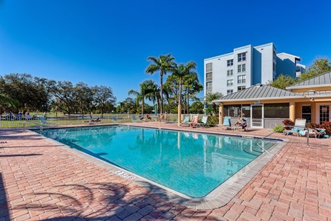 A large swimming pool surrounded by a brick patio with people relaxing in the background.