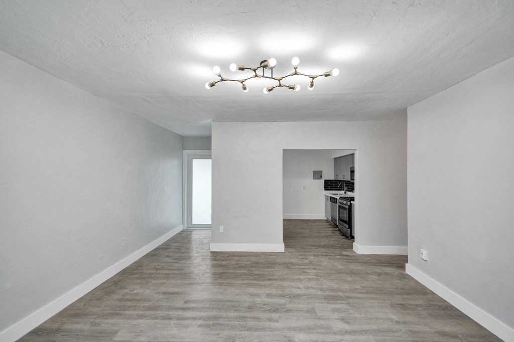 A large empty room with a chandelier and a kitchen in the background.