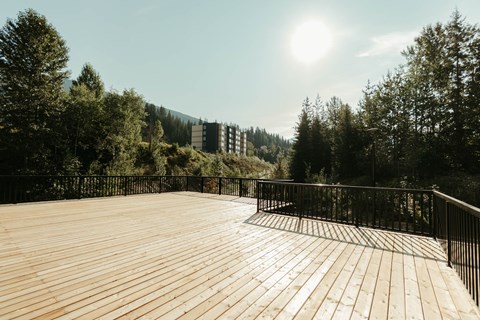 A wooden deck with railings overlooks a forest and a building in the distance.
