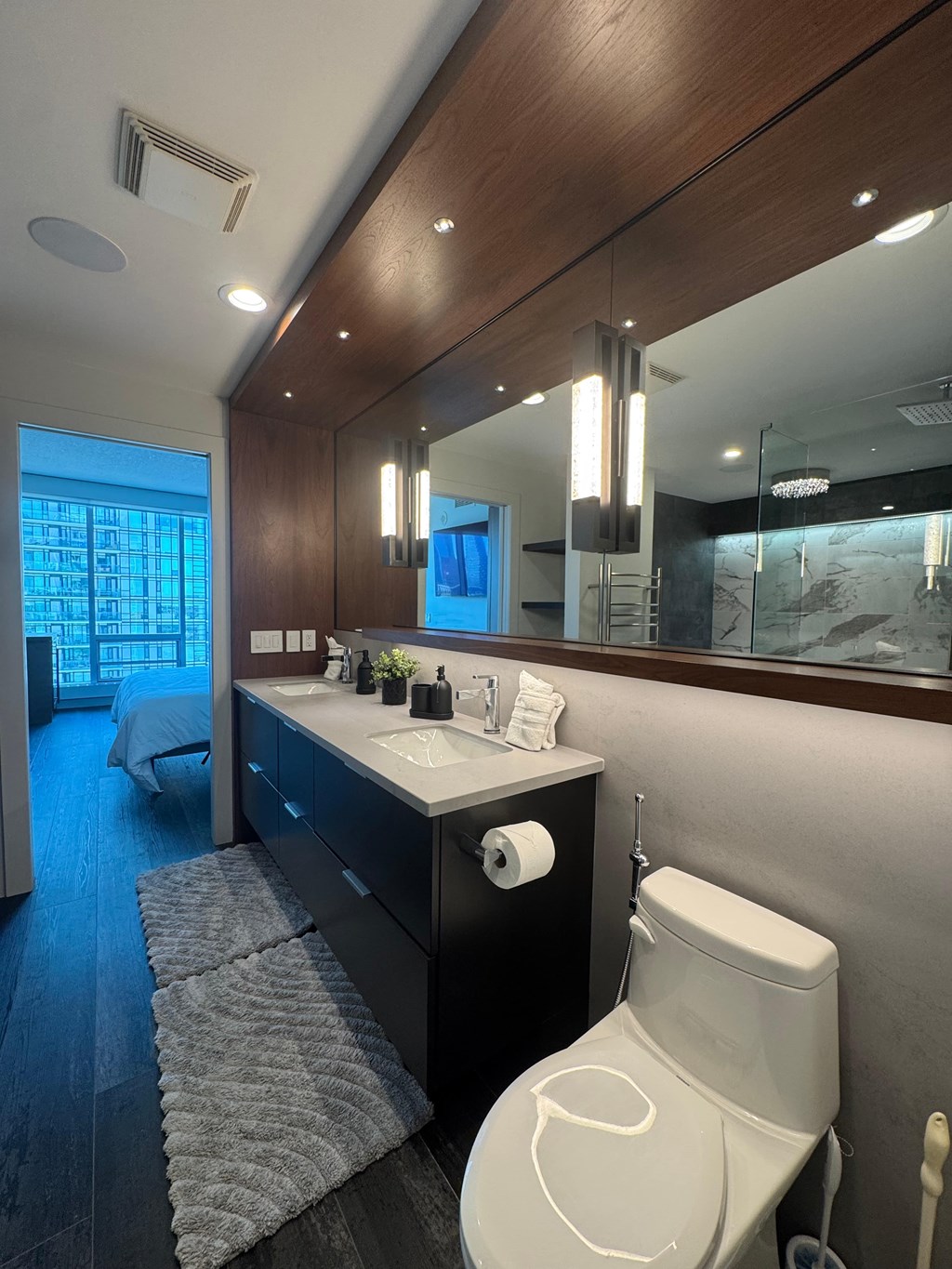 A white toilet sits in a modern bathroom with a large mirror and a bed in the background.