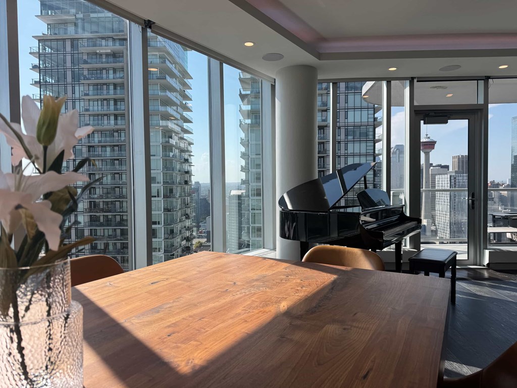 A room with a wooden table and a piano in front of a large window with a city view.