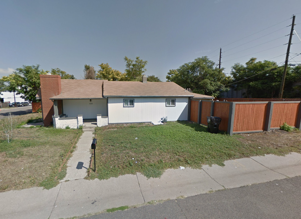 A house with a white exterior and a brown roof is situated on a street corner.