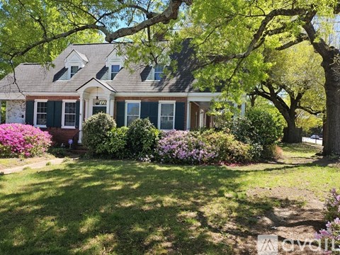A house with green shutters and a red door is surrounded by trees and flowers.