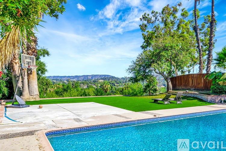 A pool surrounded by a green lawn and trees with a basketball hoop.