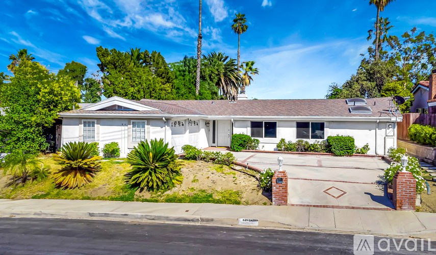 A house with a white exterior and a brown roof is surrounded by greenery and trees.