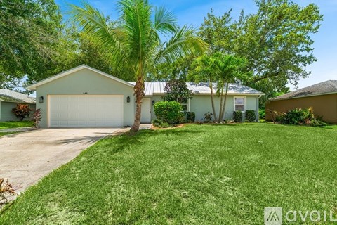 A house with a white garage door is surrounded by greenery and palm trees.