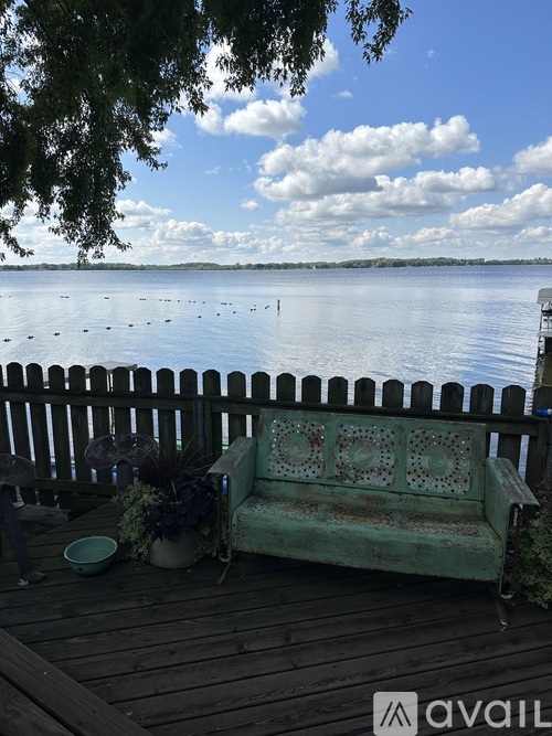 A wooden bench with a cushion sits on a wooden deck overlooking a lake.