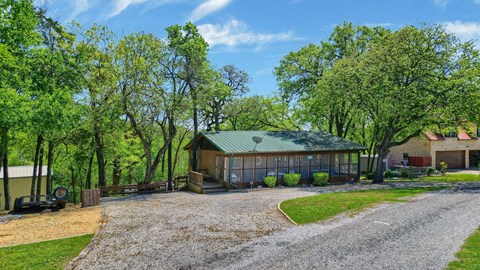 A house with a green roof is surrounded by trees and has a gravel driveway leading to the front door.