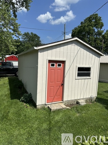 A small white shed with a red door is situated in a grassy area.