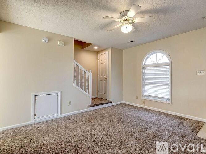 A room with a carpeted floor, a ceiling fan, and a staircase.