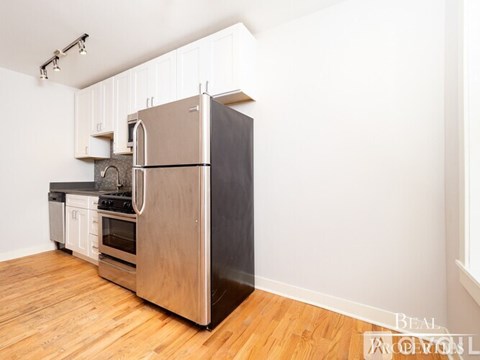 A stainless steel refrigerator stands in a kitchen with wooden floors and white walls.
