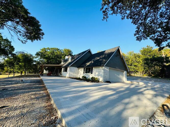 A white house with a black roof is surrounded by trees and has a gravel driveway leading to the garage.
