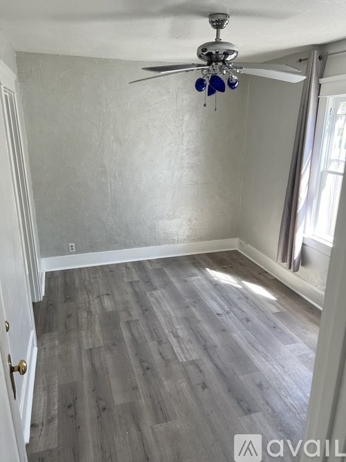 A room with a ceiling fan and light, hardwood floors, and a window with curtains.