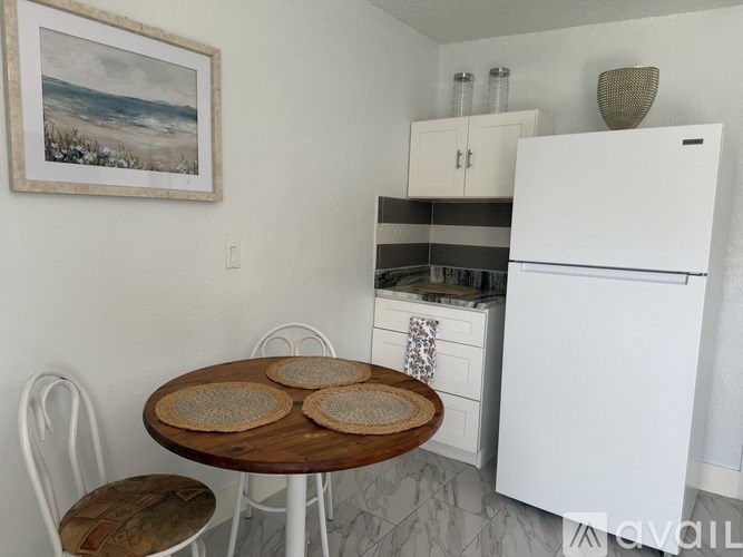 A kitchen with a white refrigerator, a wooden table with three round woven placemats, and white chairs.