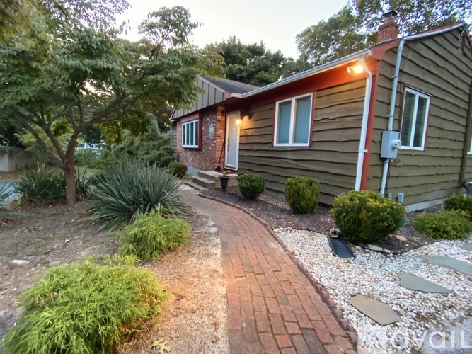 A house with a brown siding and a red roof is surrounded by a garden with a brick pathway.