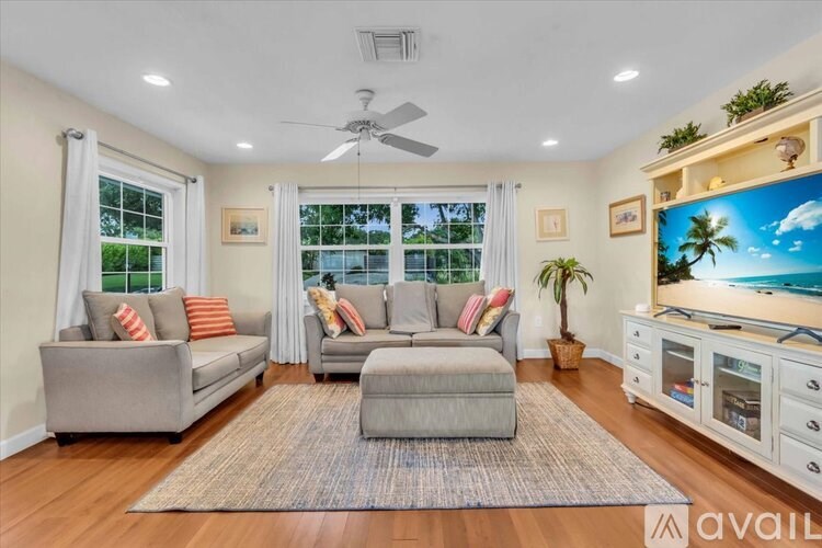 A living room with a grey couch, a rug, a ceiling fan, and a painting of a beach scene on the wall.