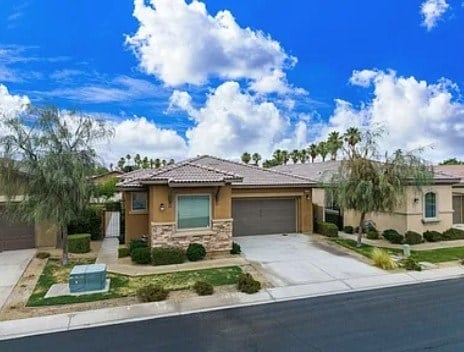 A house with a brown roof and a grey garage door is surrounded by green shrubs and trees.