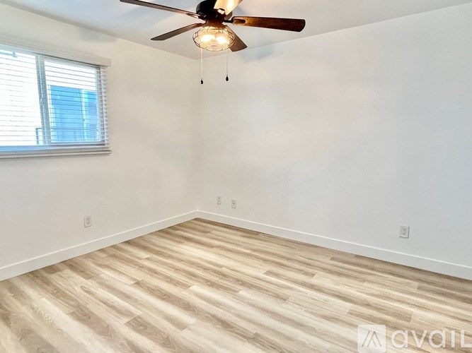 A room with a ceiling fan and light fixture, wooden flooring, and a window with blinds.