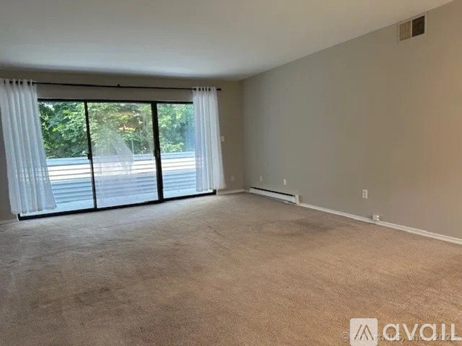 A spacious room with a carpeted floor, large windows with white curtains, and a view of a street outside.