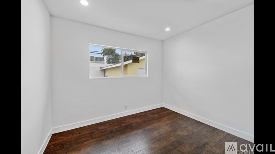 A room with wooden flooring and white walls, with a window showing a view of a building and trees.