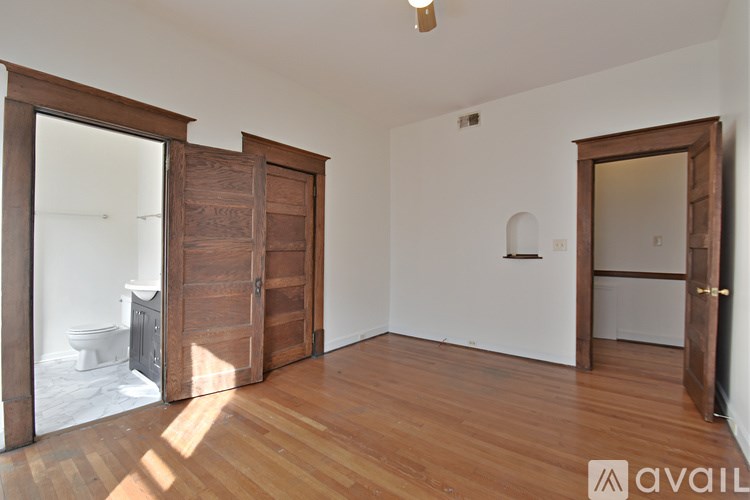A room with wooden floors and white walls, with a toilet visible through the open door.