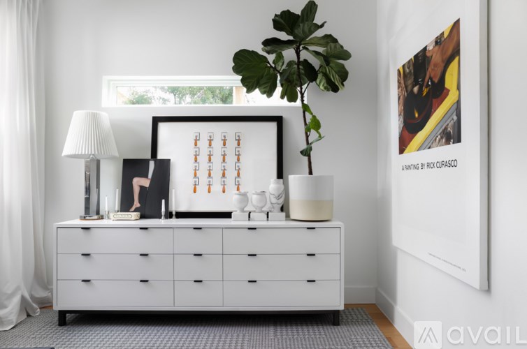 A white dresser with a plant on top and a framed picture on the wall.