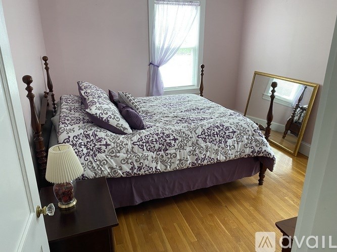 A bed with a purple and white comforter sits in a room with wooden floors and a window with white curtains.