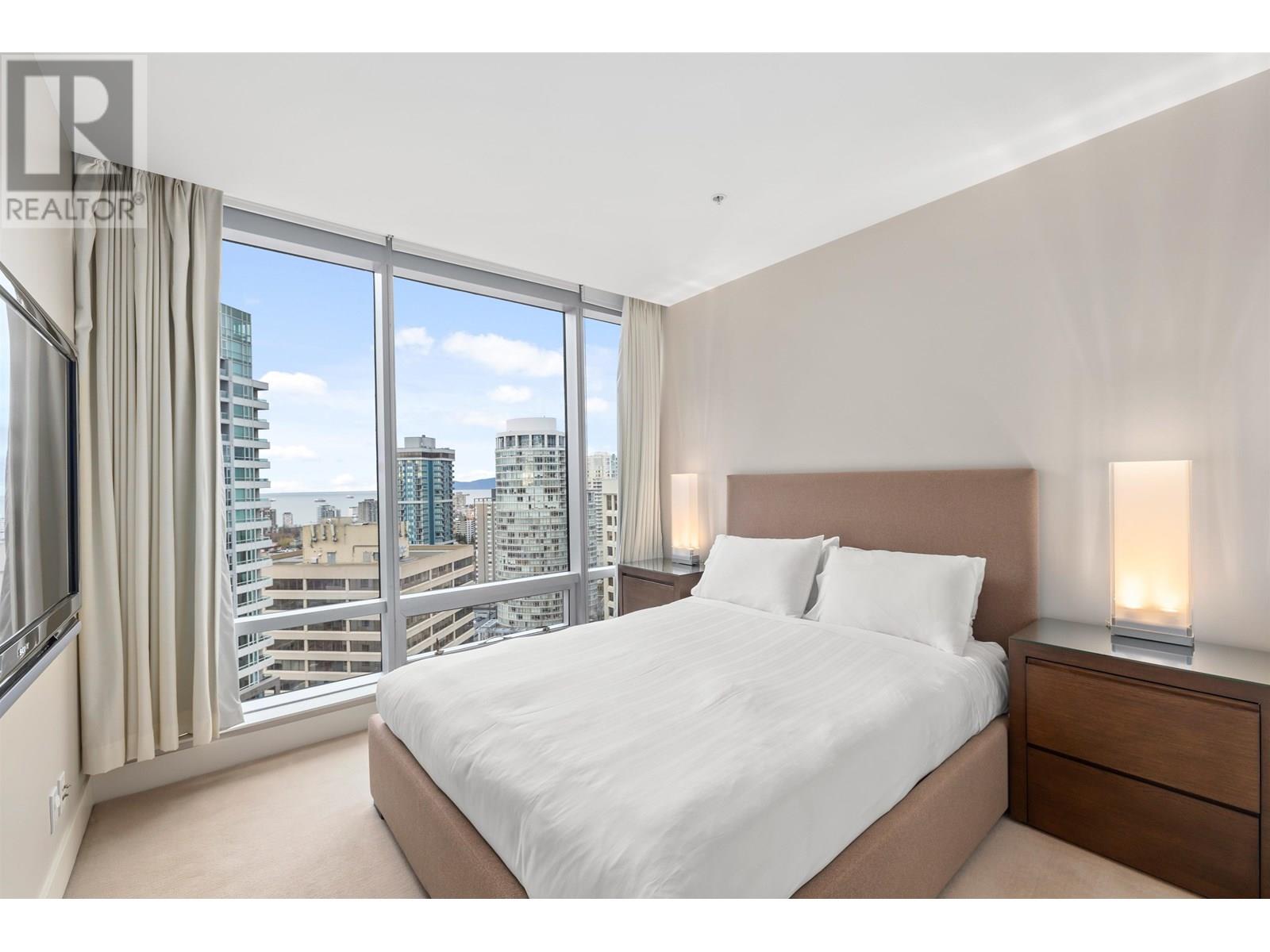 A bedroom with a large bed, a window with a view of the city, and a nightstand with a lamp.